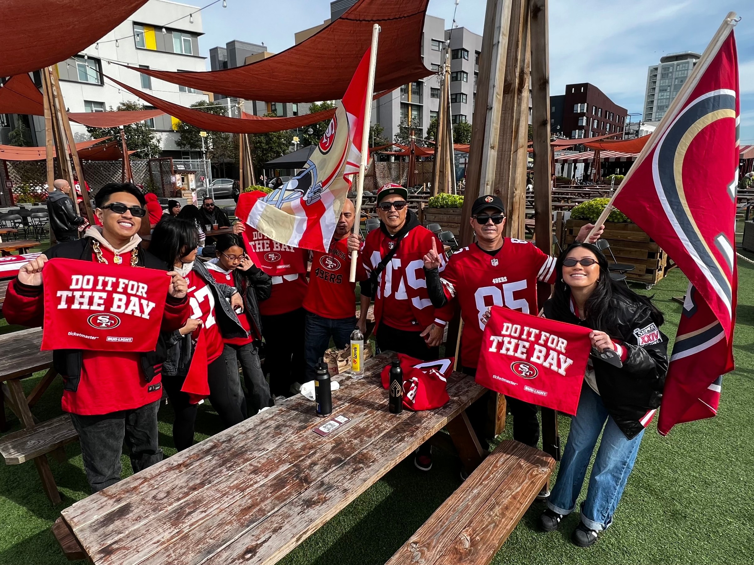 Group of people in 49ers jerseys holding flags and signs, gathered outdoors by picnic tables.