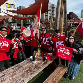 Group of people in 49ers jerseys holding flags and signs, gathered outdoors by picnic tables.