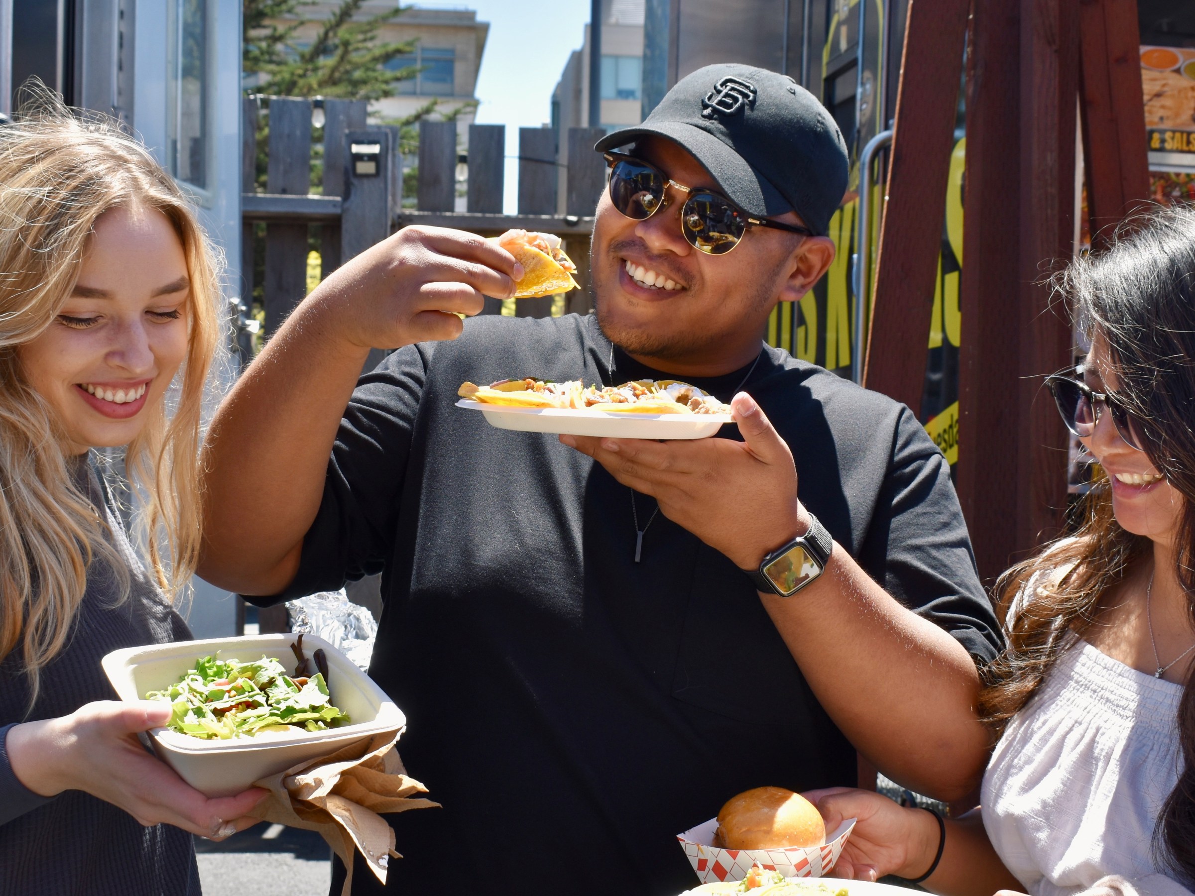 Three people smiling and holding plates of food at an outdoor food truck event.