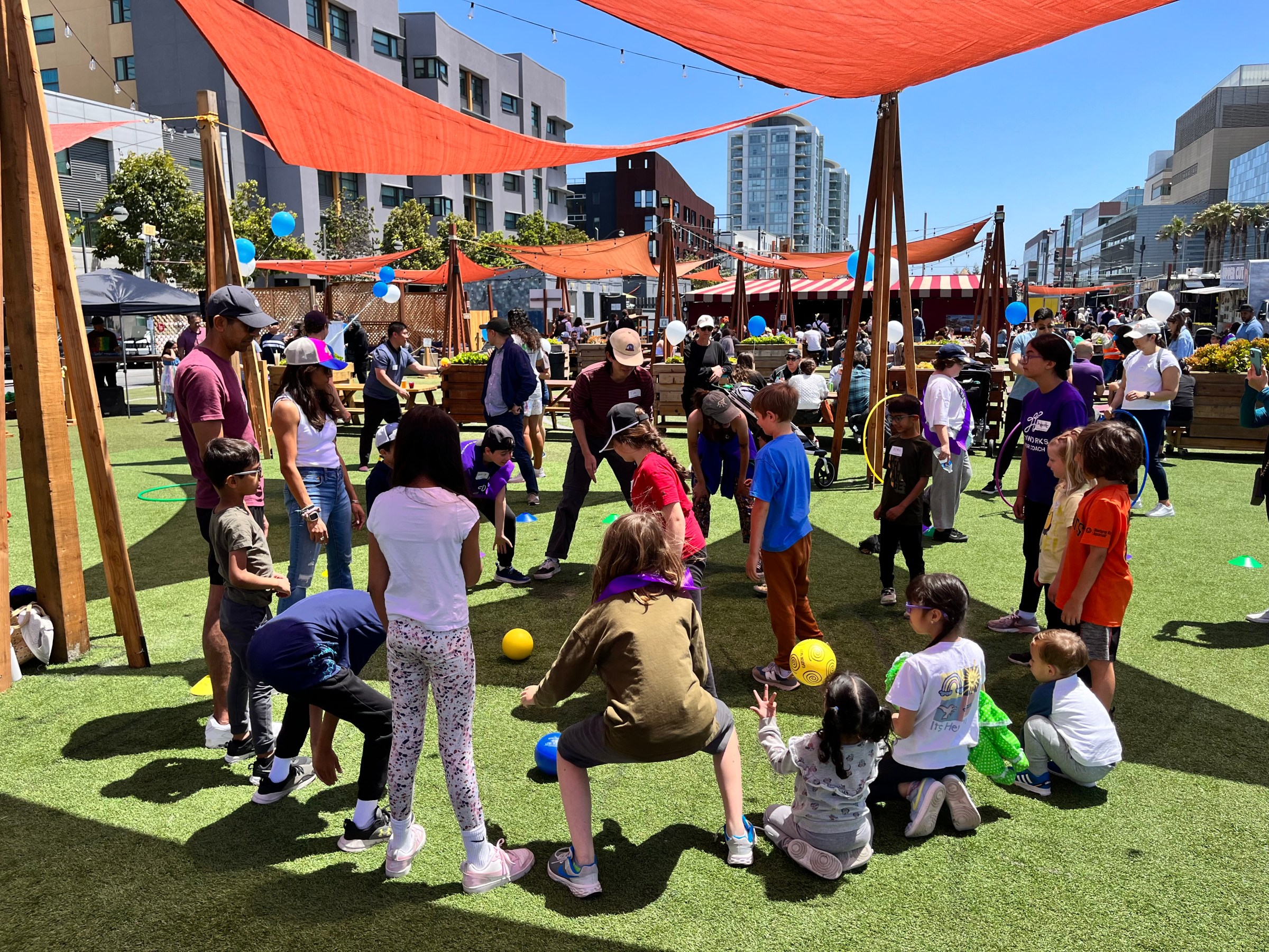 Children and adults play games outdoors on artificial grass under orange canopies.