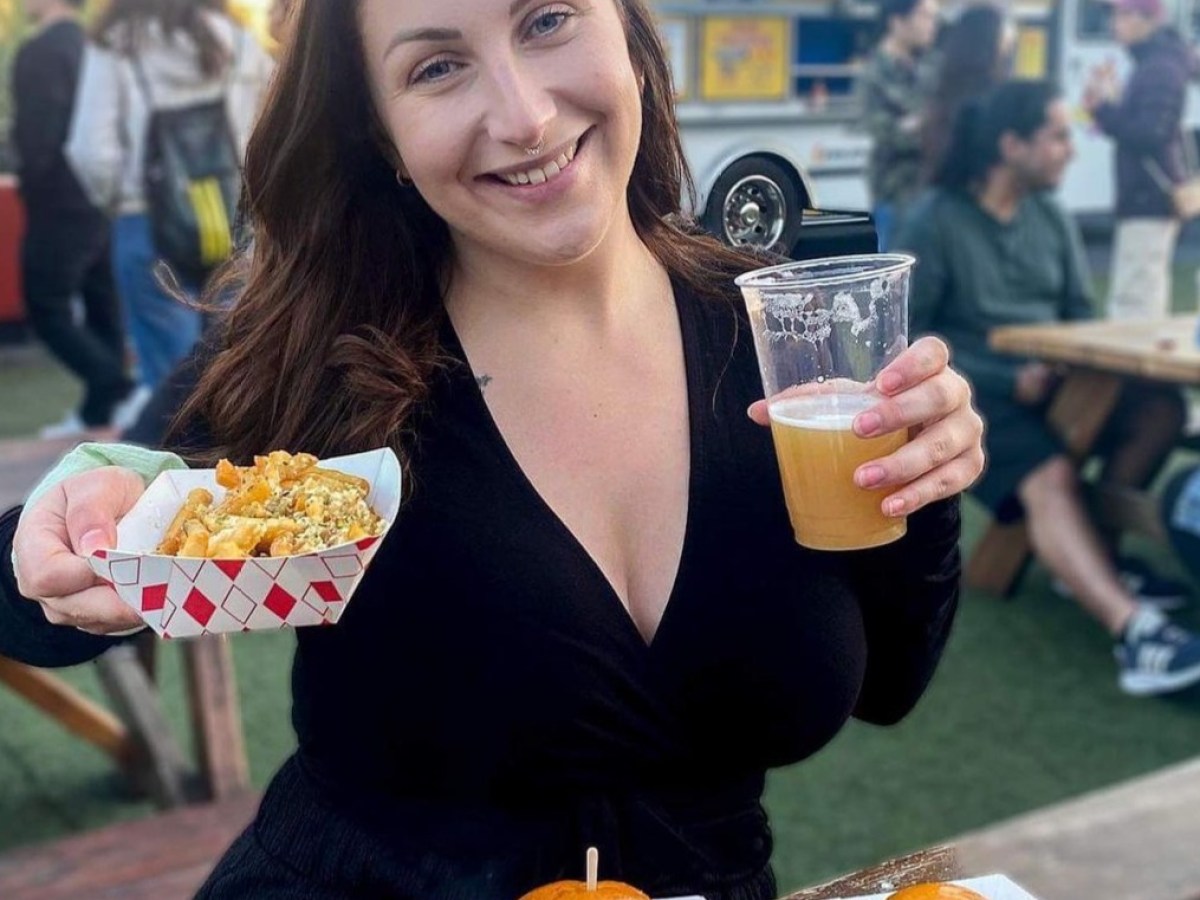 Woman holding fries and beer with sandwiches in front, food truck in background.