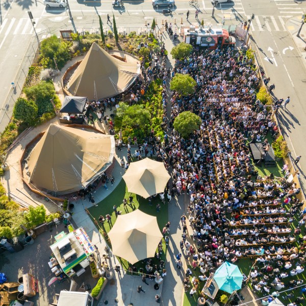 Aerial view of a large crowd gathered outdoors near tents and food trucks, adjacent to a city intersection.