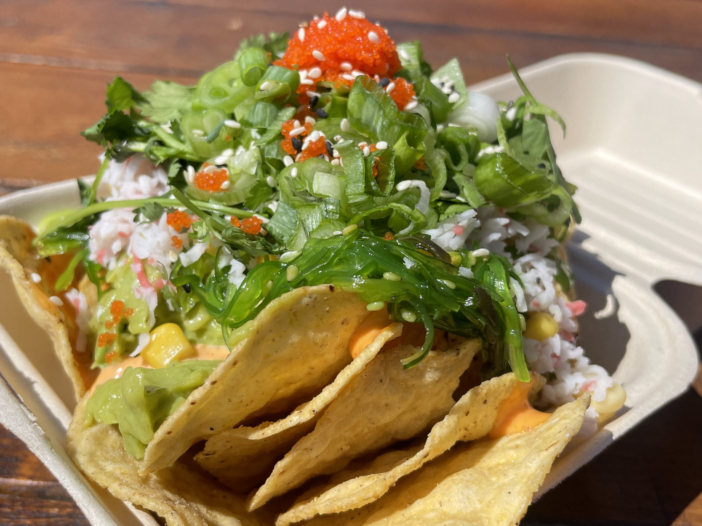 Nachos with guacamole, green onions, and red seasoning in a cardboard tray on a wooden table.