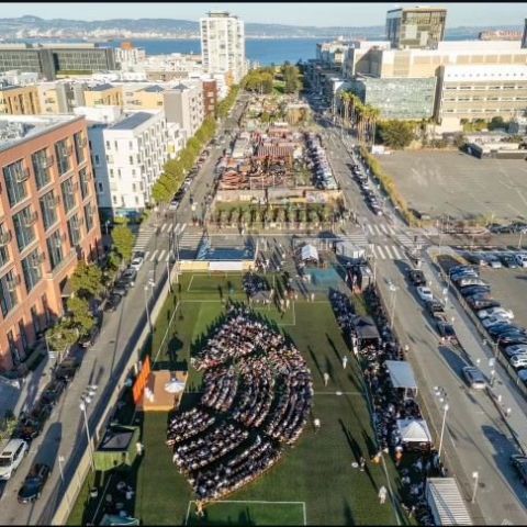 Aerial view of a city event with a large audience seated in a park, surrounded by buildings and streets.