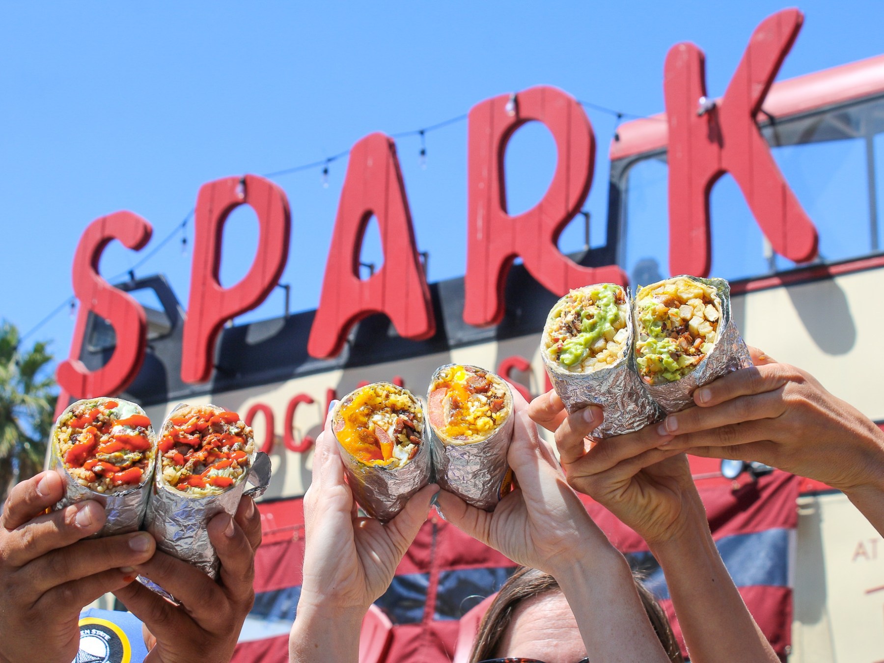 Hands holding various foil-wrapped burritos in front of large red 'SPARK' sign under blue sky.