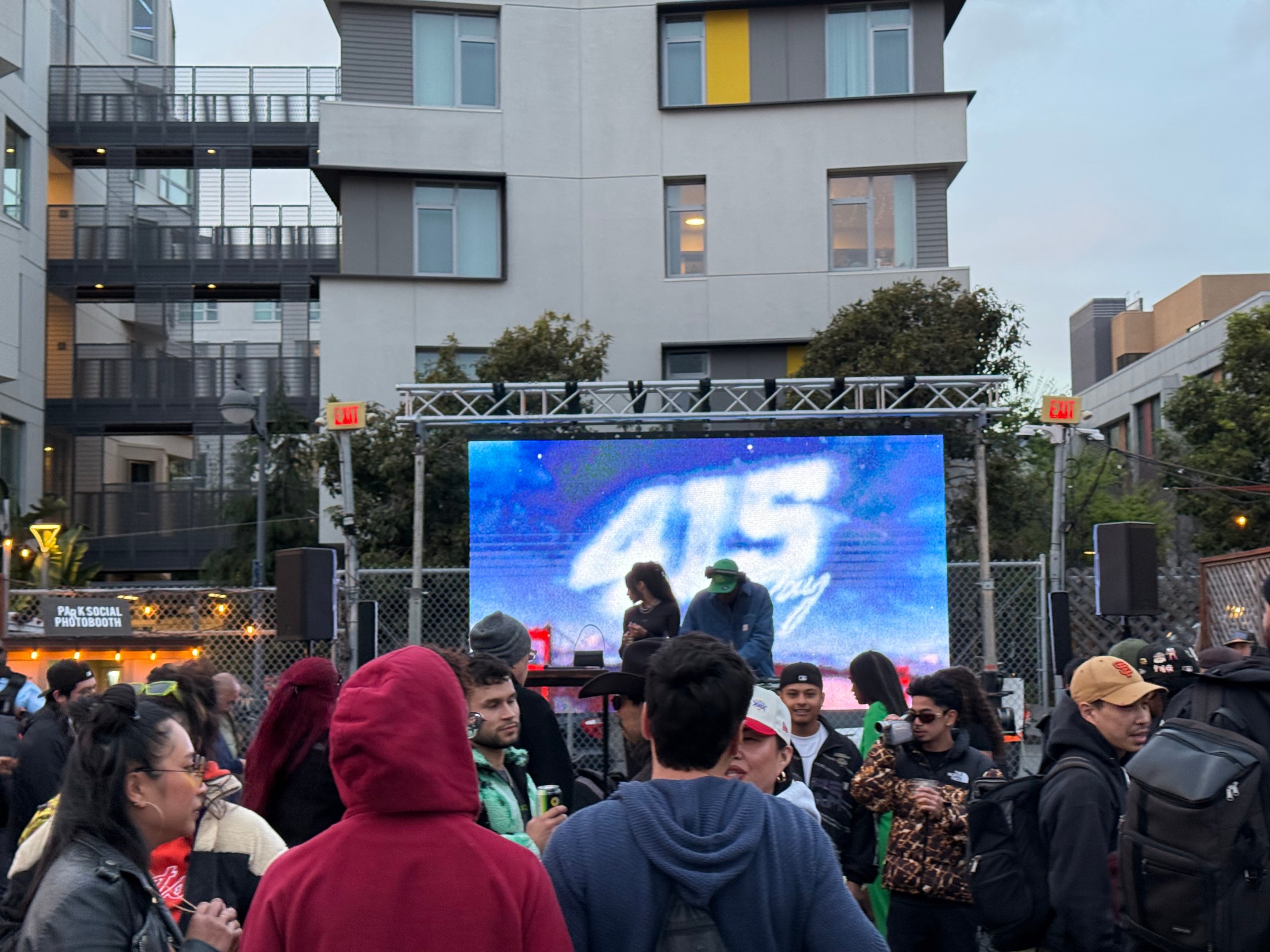 Outdoor gathering with people in front of stage and screen in urban setting.