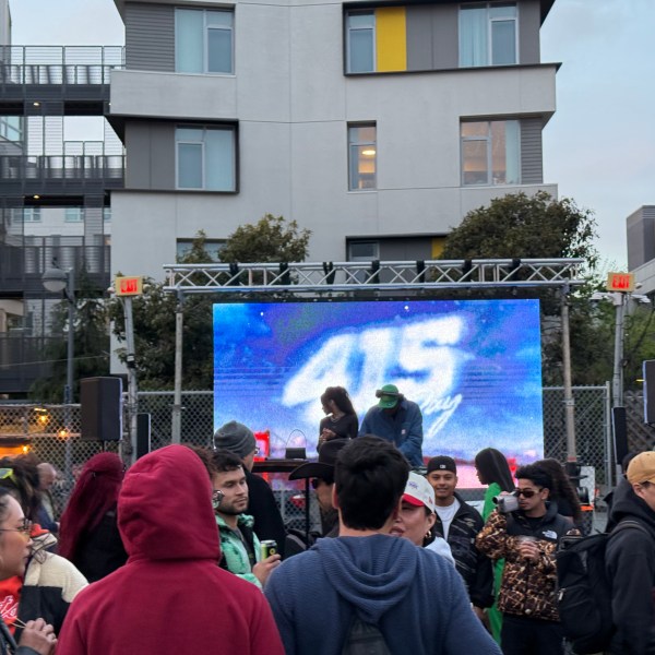 Outdoor gathering with people in front of stage and screen in urban setting.