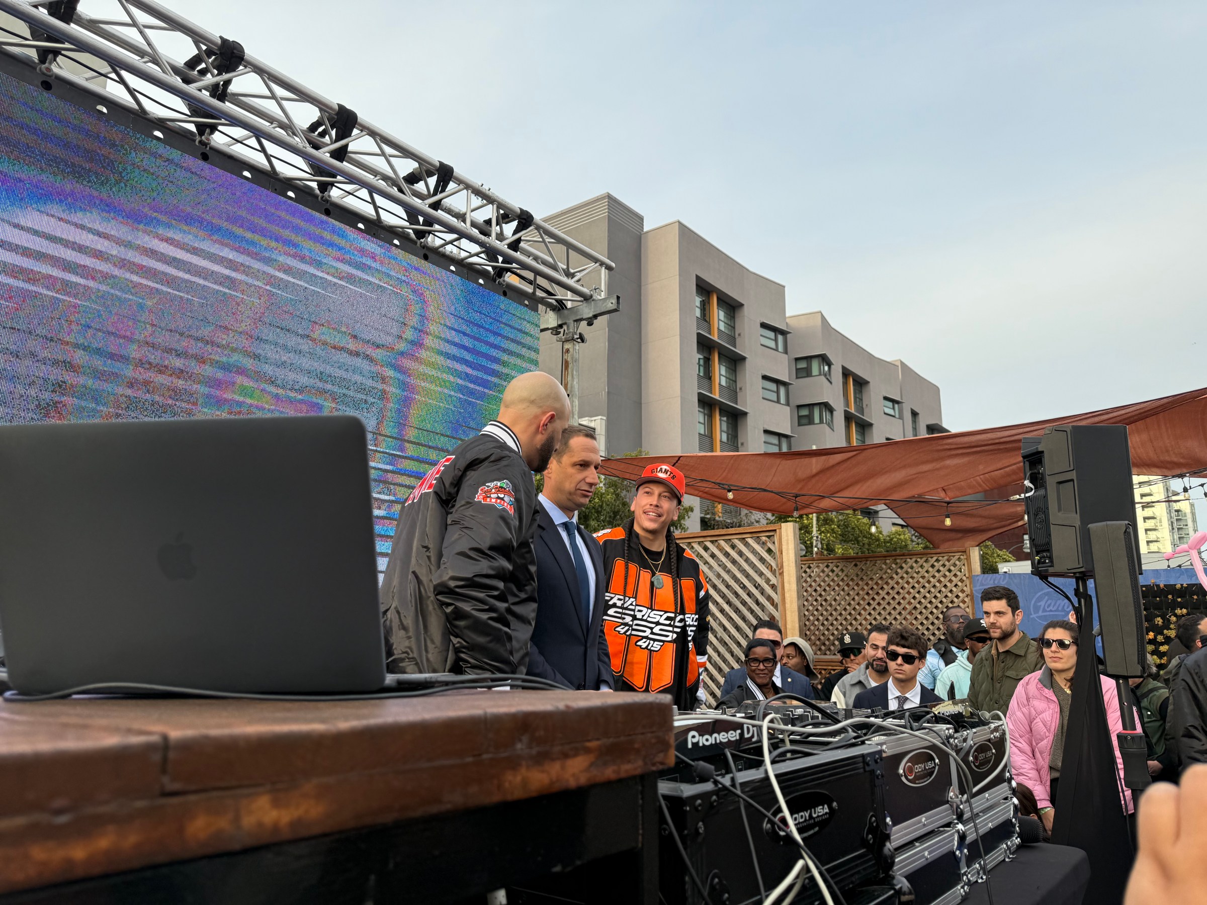 DJ booth with crowd, large screen, and buildings in the background.