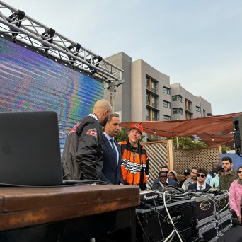 DJ booth with crowd, large screen, and buildings in the background.