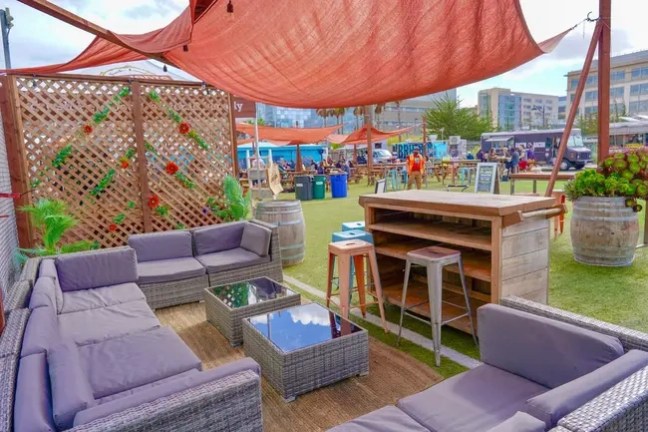 Outdoor lounge with sofas, wooden bar, stools, and sunshade canopy on grass.