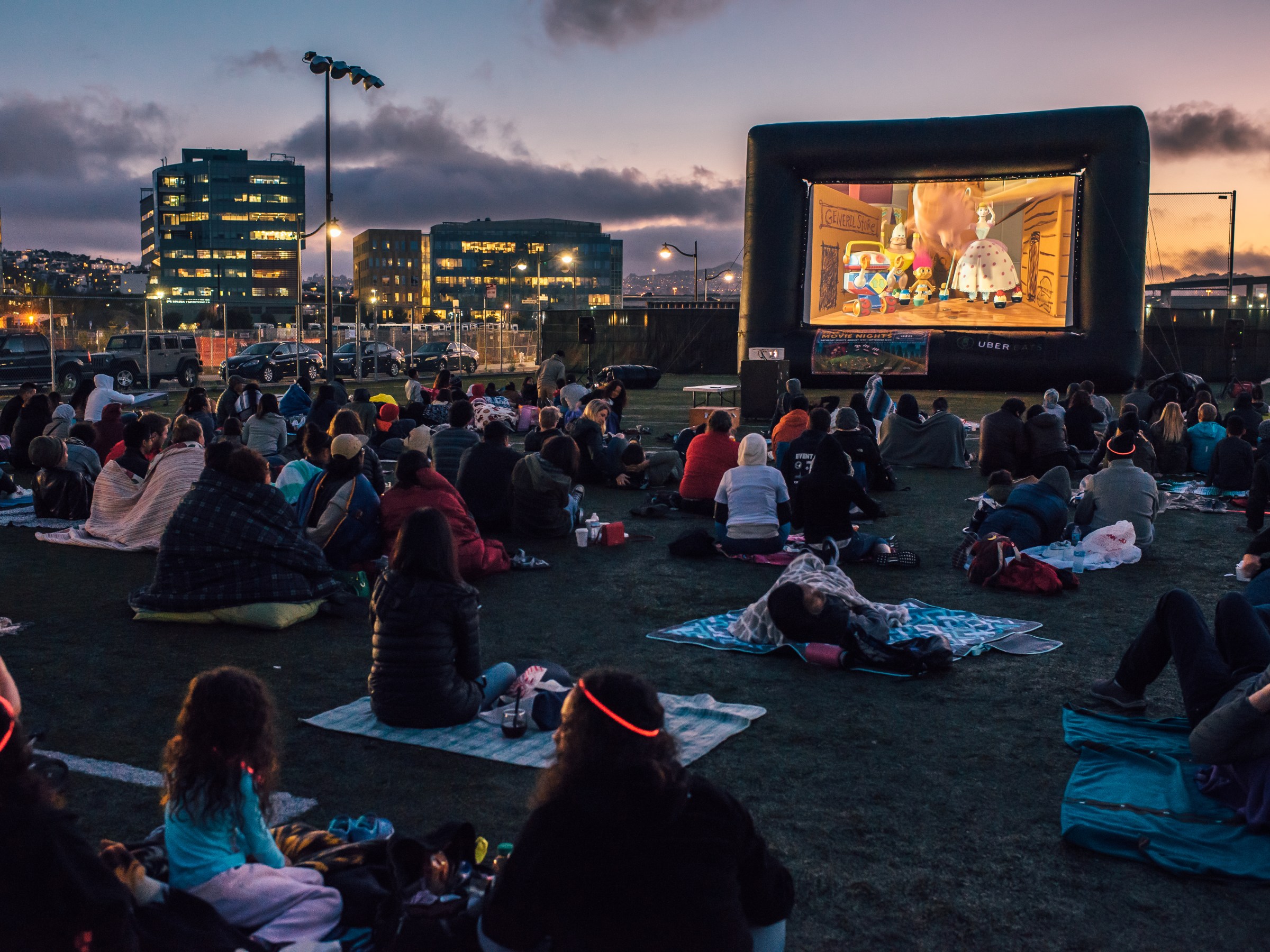 People watching an outdoor movie on a large screen at night, with city lights in the background.