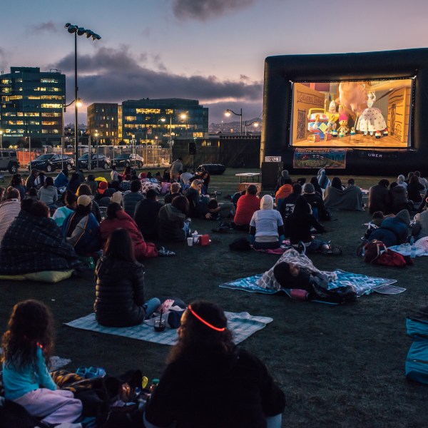 People watching an outdoor movie on a large screen at night, with city lights in the background.
