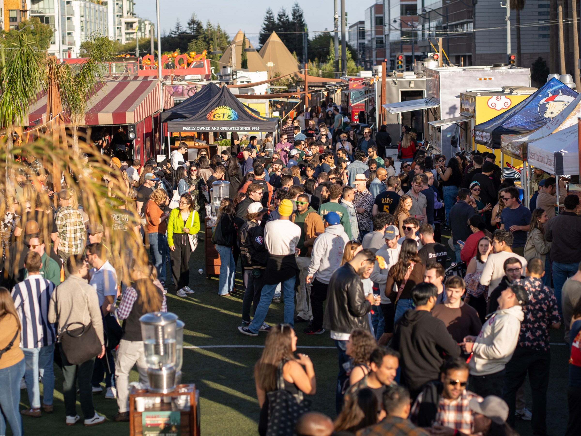 Crowded outdoor market with people mingling among food stalls and tents on a sunny day.