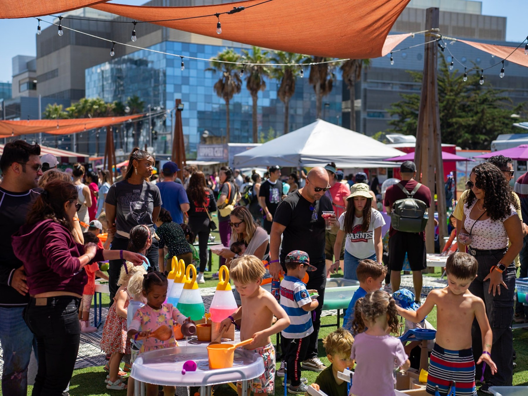 Crowd at outdoor market with children playing at activity tables under shade sails.