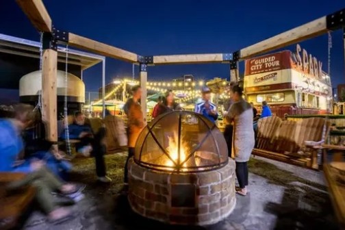 People gathered around a fire pit with swings at night, outdoor venue with lights and a food truck.