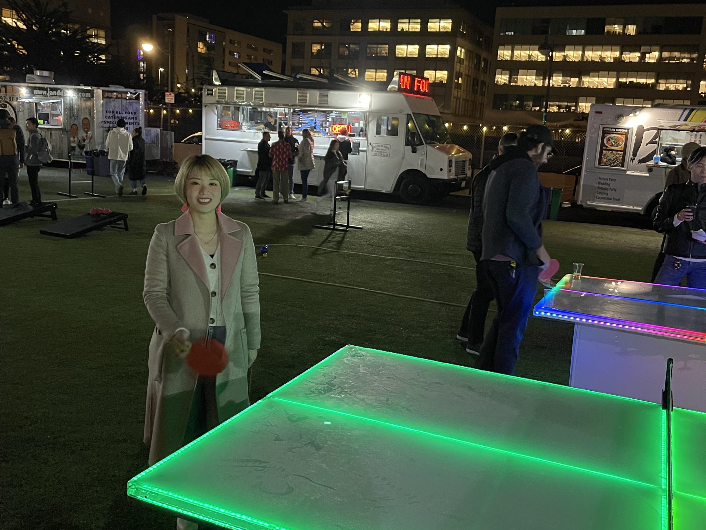 Person playing ping pong at night by food trucks and illuminated tables.