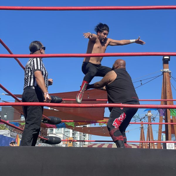 Wrestler in mid-air above opponent in a wrestling ring with referee nearby.