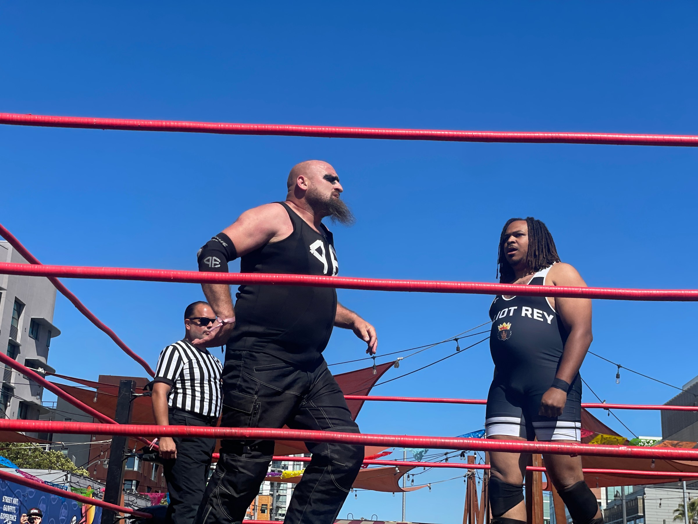 Two wrestlers face off in a ring outdoors, with a referee nearby.