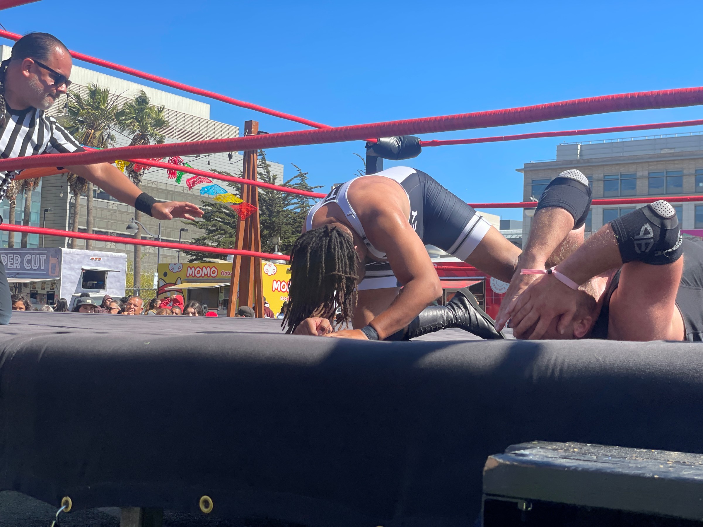 Wrestler pins opponent in outdoor ring with referee nearby, clear blue sky.