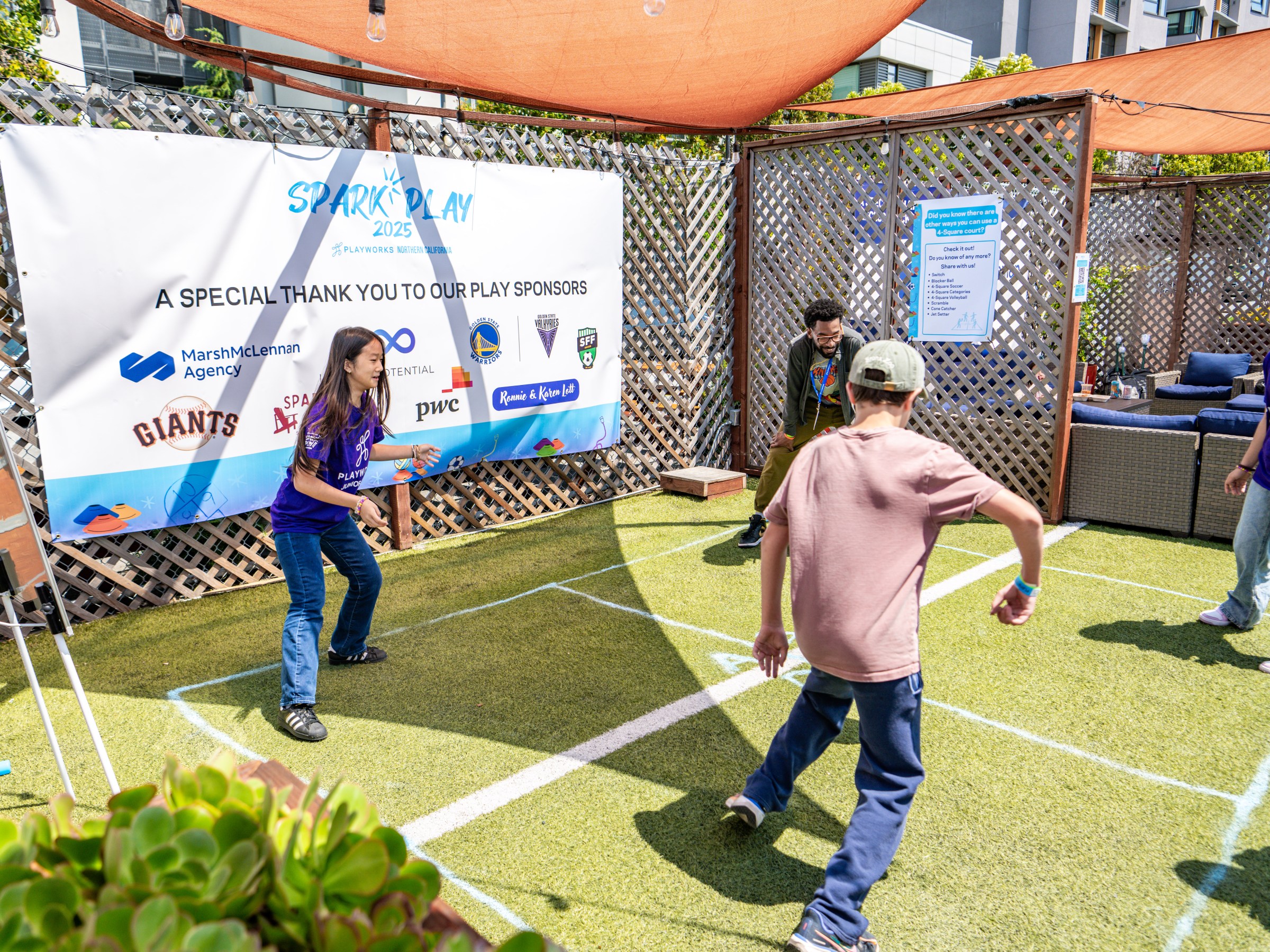 Four people playing a ball game on a grass court under an awning, near a sign thanking sponsors.