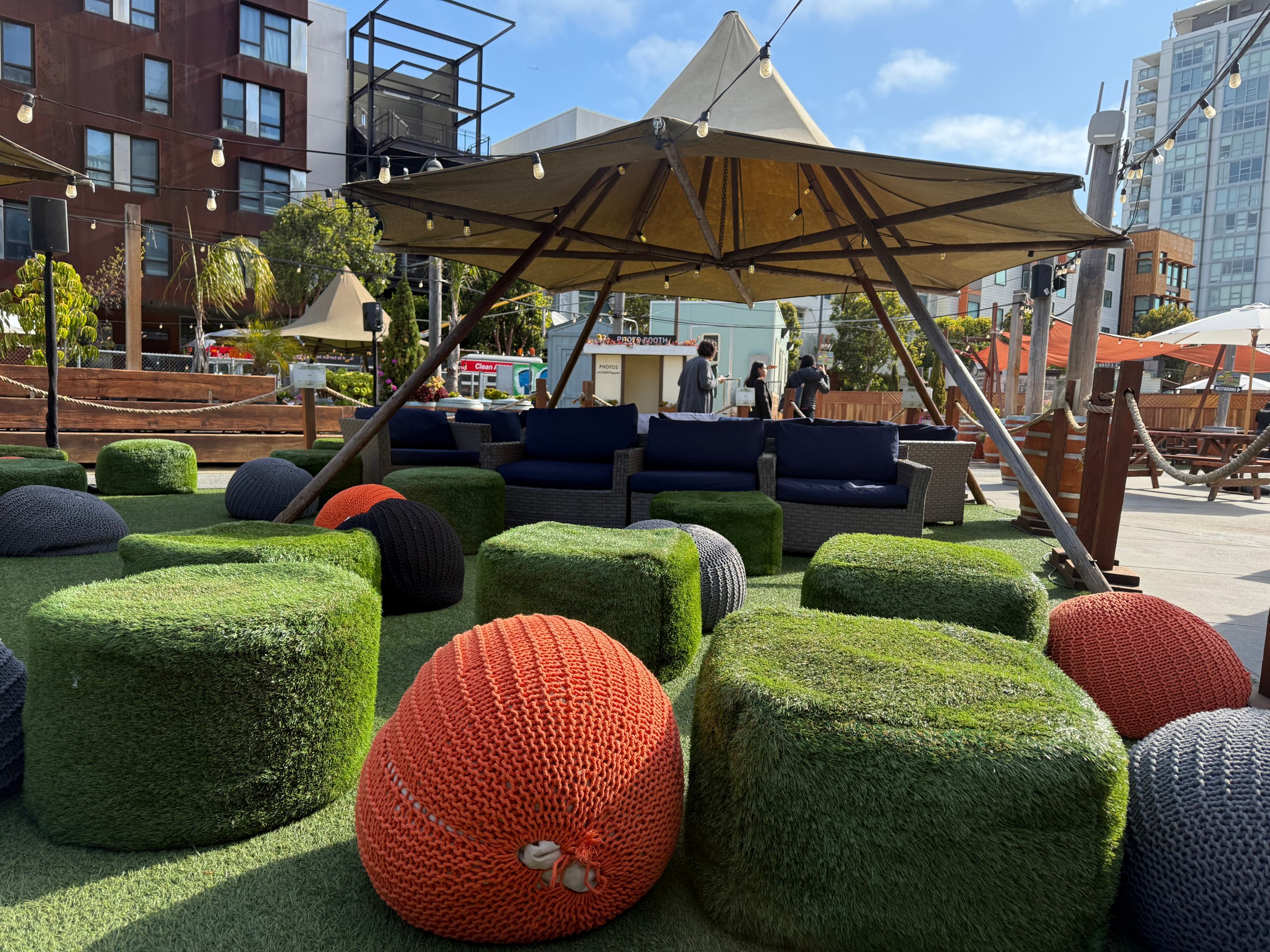 Outdoor seating area with grass-covered and knitted poufs under a large tent.
