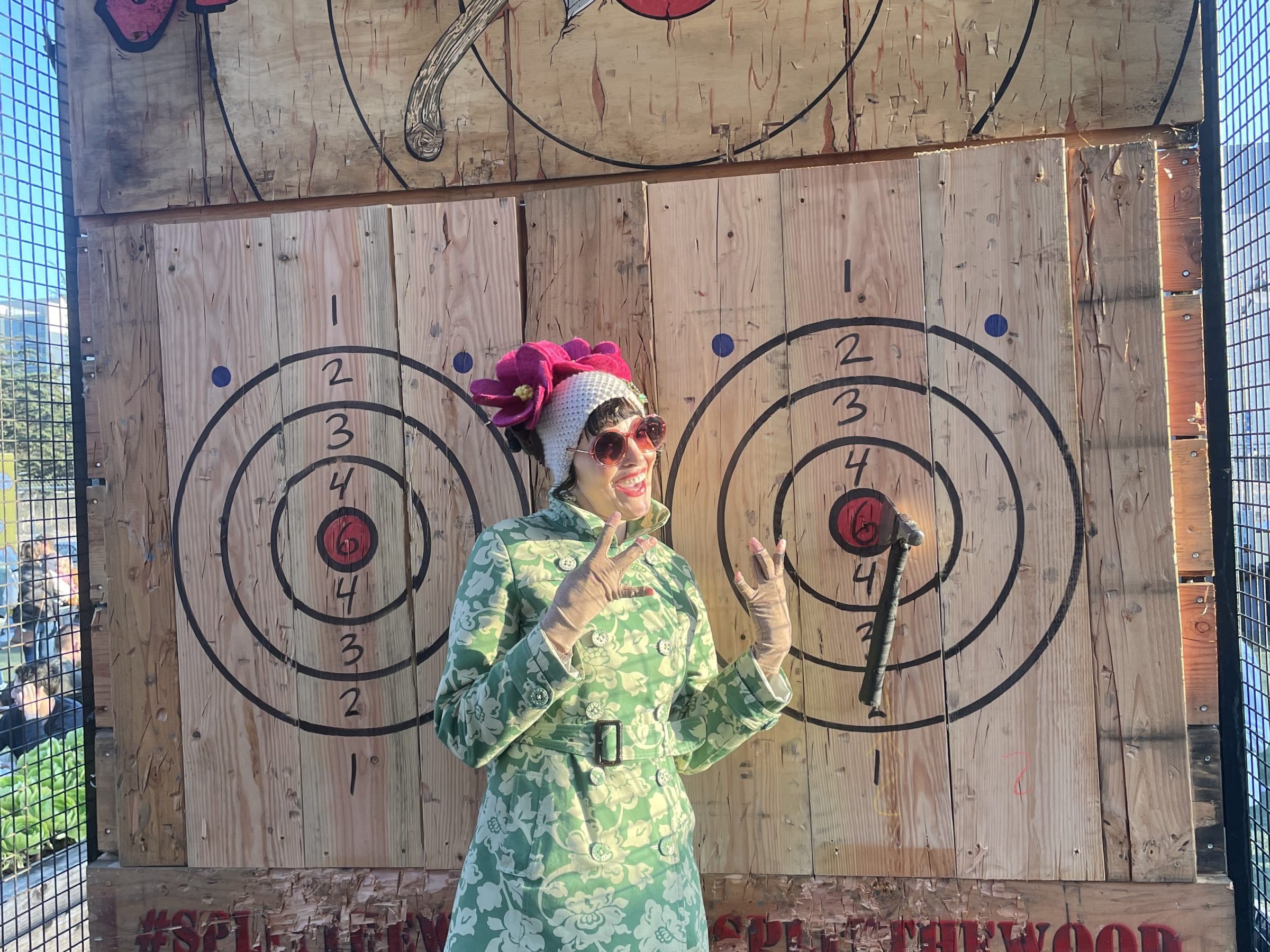 Person in floral coat and hat poses in front of axe-throwing target at 'Split the Wood' venue.