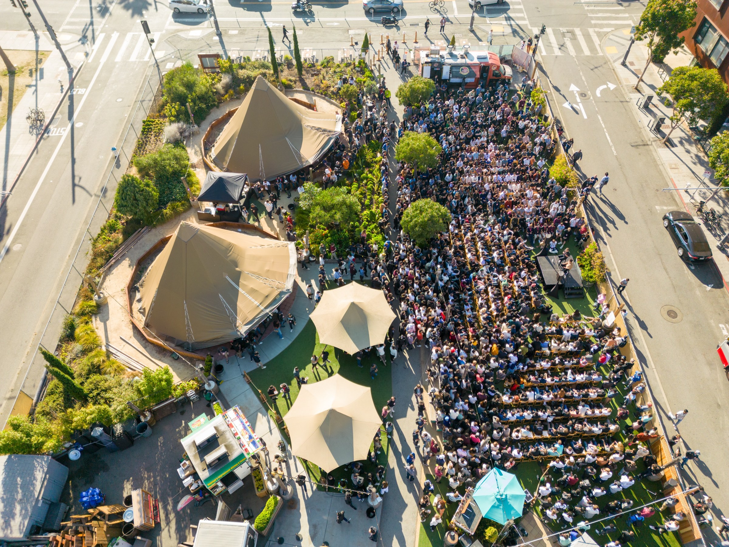 Aerial view of a large outdoor gathering with tents and people seated on benches.