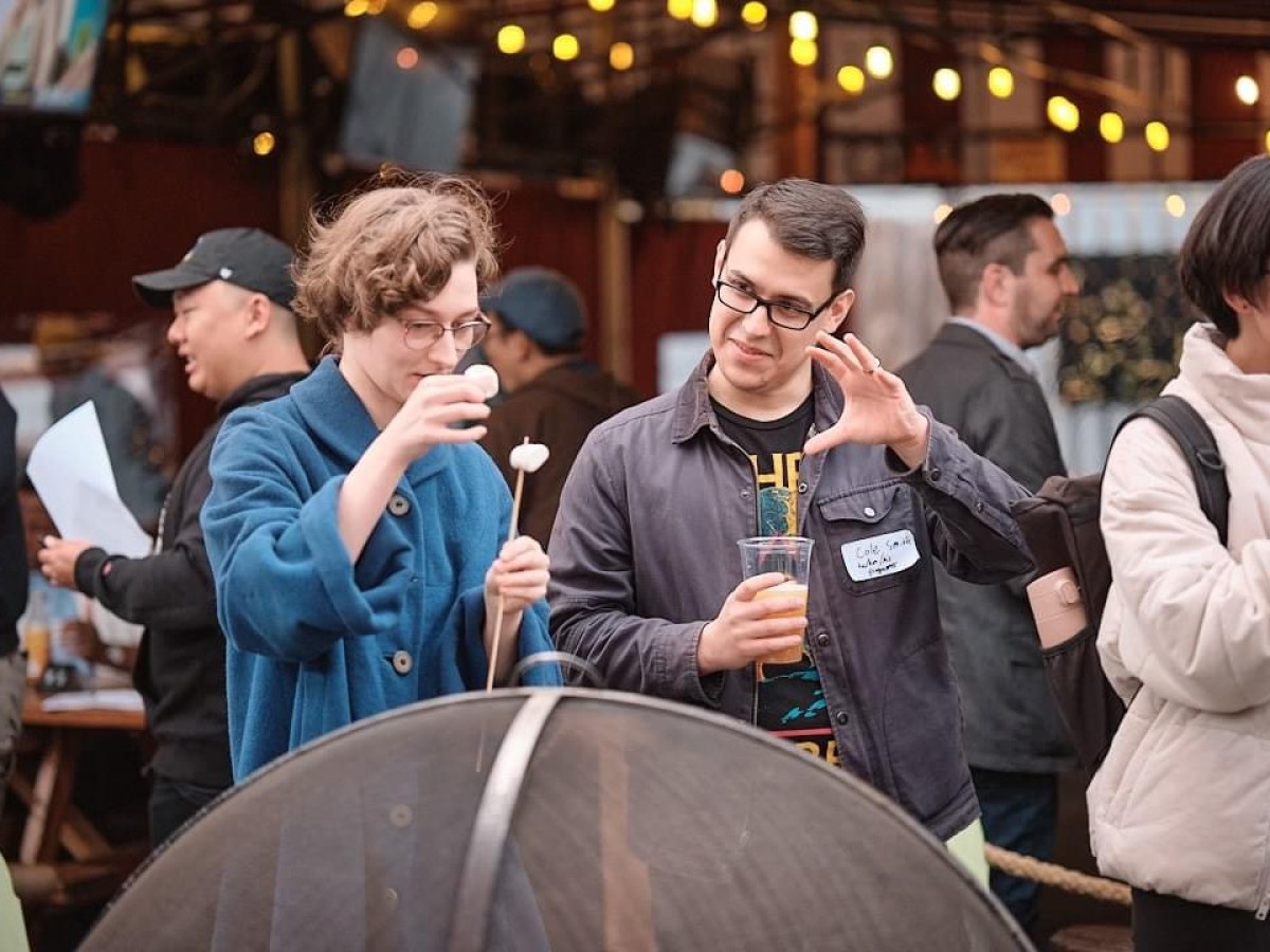 Two people at a gathering, one holding a marshmallow on a stick, the other with a drink, in an outdoor setting.