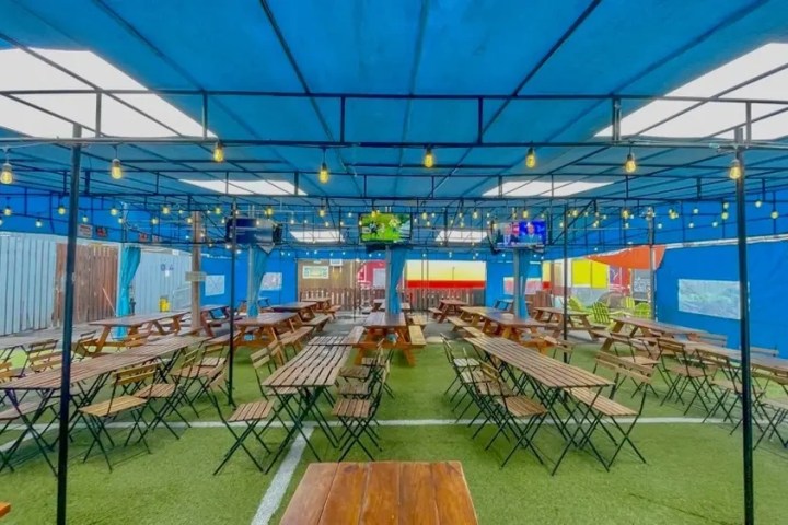 Outdoor seating area with wooden tables, chairs, string lights, and a blue canopy on artificial grass.