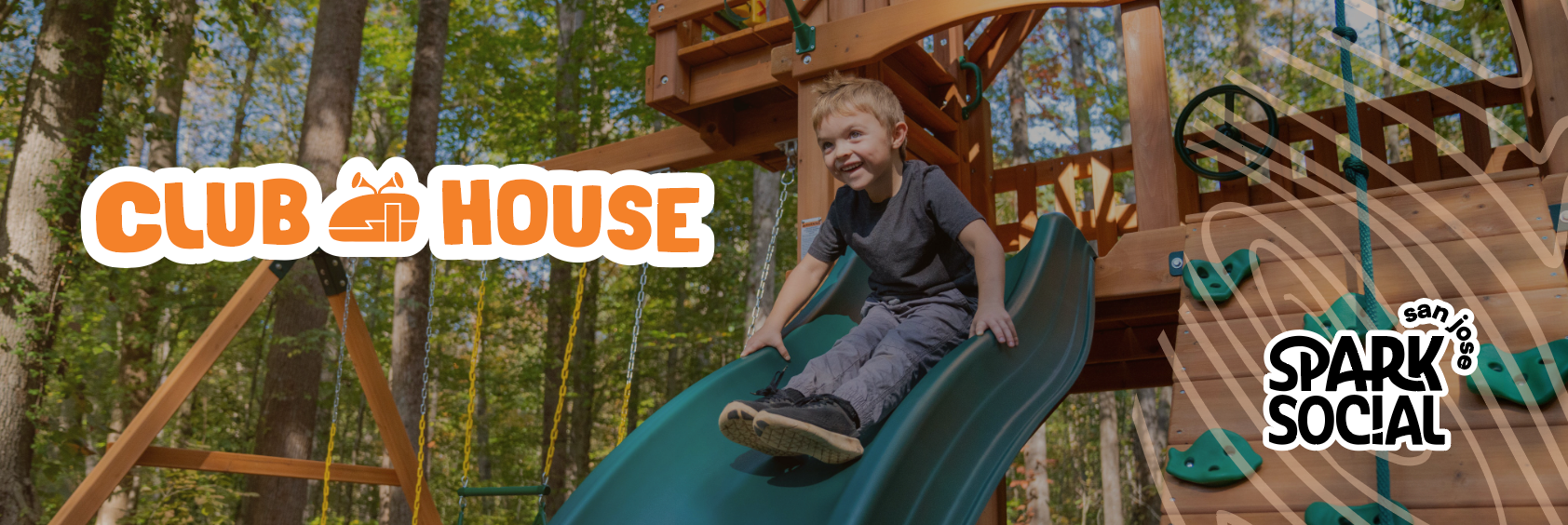 Child sliding down a green slide in a wooden play structure in a forested area.