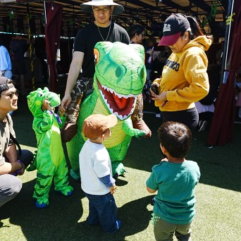 People and kids in dinosaur costumes interacting at an outdoor event.