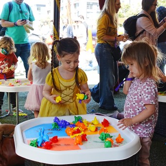 Children playing with colorful toys at a table in a crowded outdoor event.