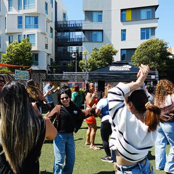 Group of people dancing outdoors on a sunny day, with modern buildings in background.