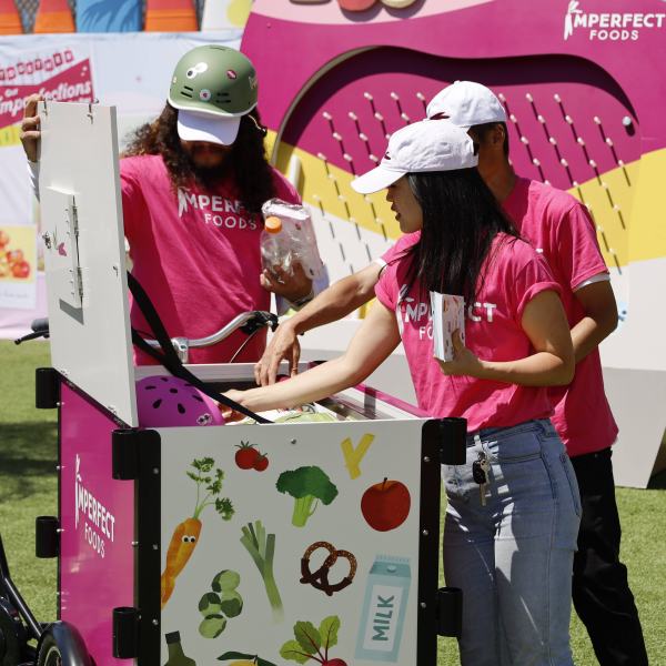Three people in pink shirts serve food from a bicycle cart on a grassy area.