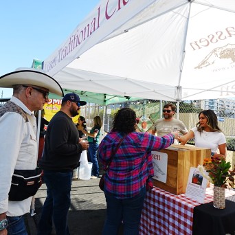 People at an outdoor market booth under a white tent, engaging with vendors.